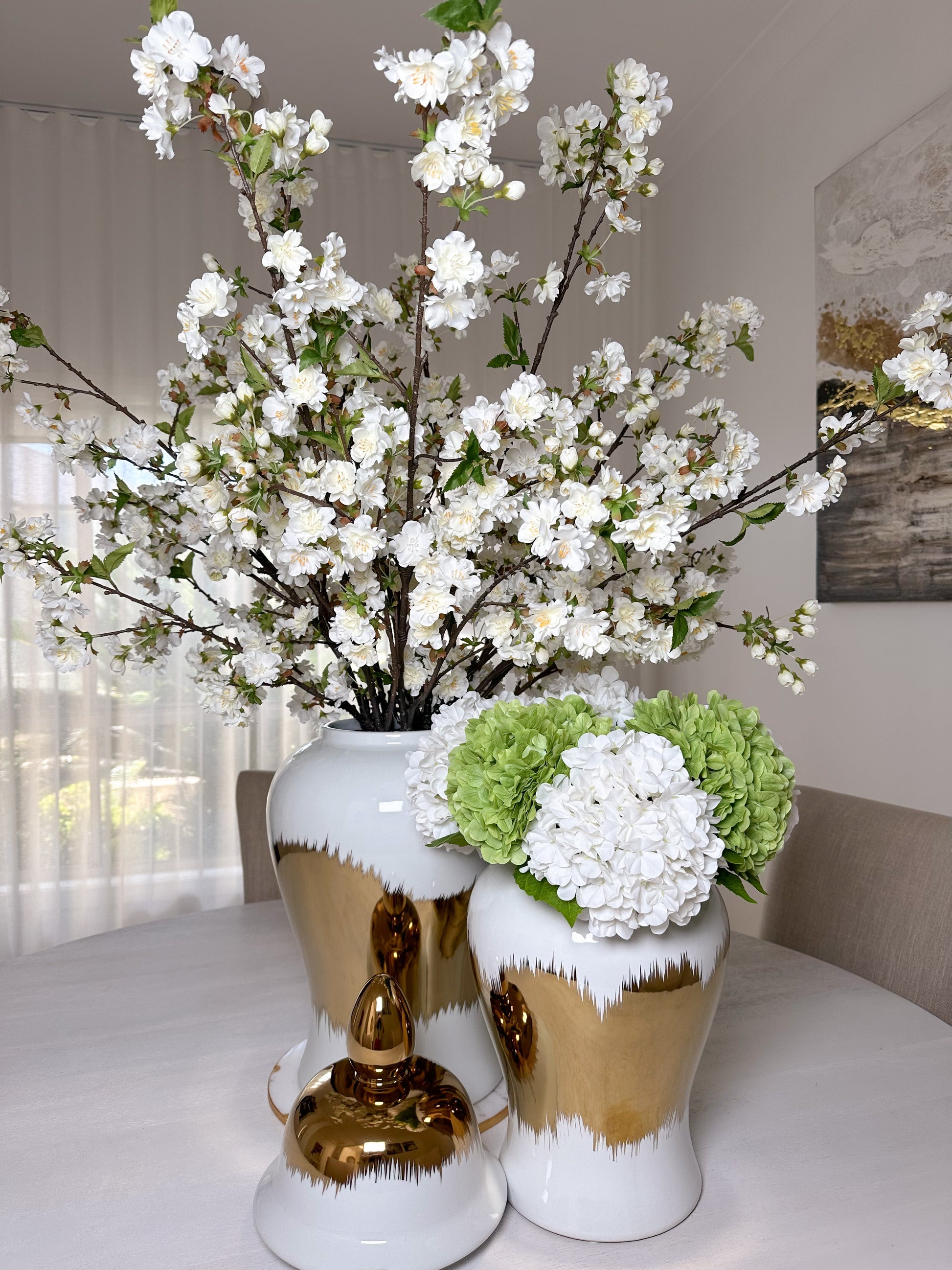 White cherry blossom stems in white and gold vase with second white and gold vase in front with white and green real touch hydrangeas on dining table front view