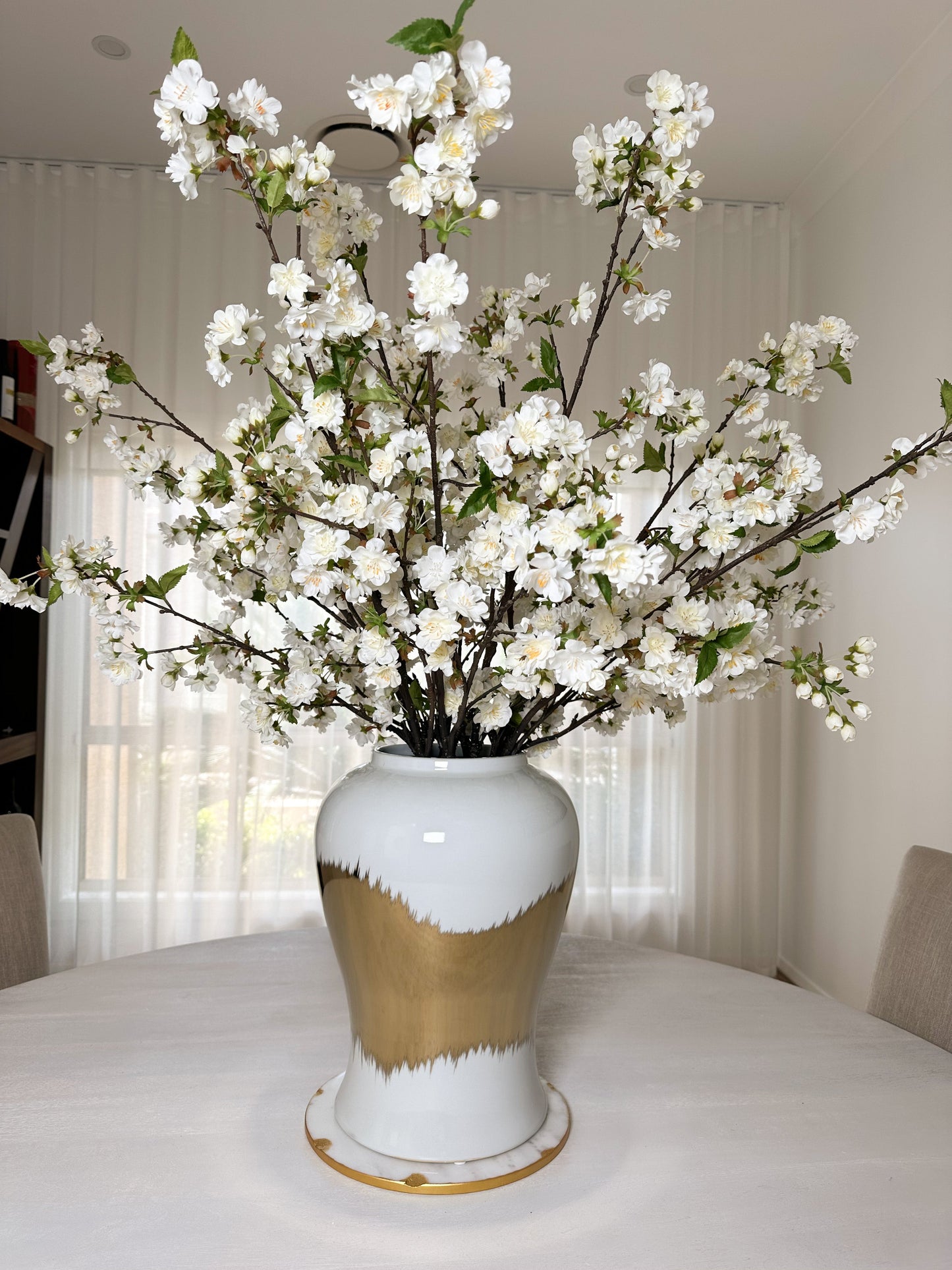 White and Gold Vase with cherry blossoms on marble board from front view on dining table. With sheer curtains in the background.