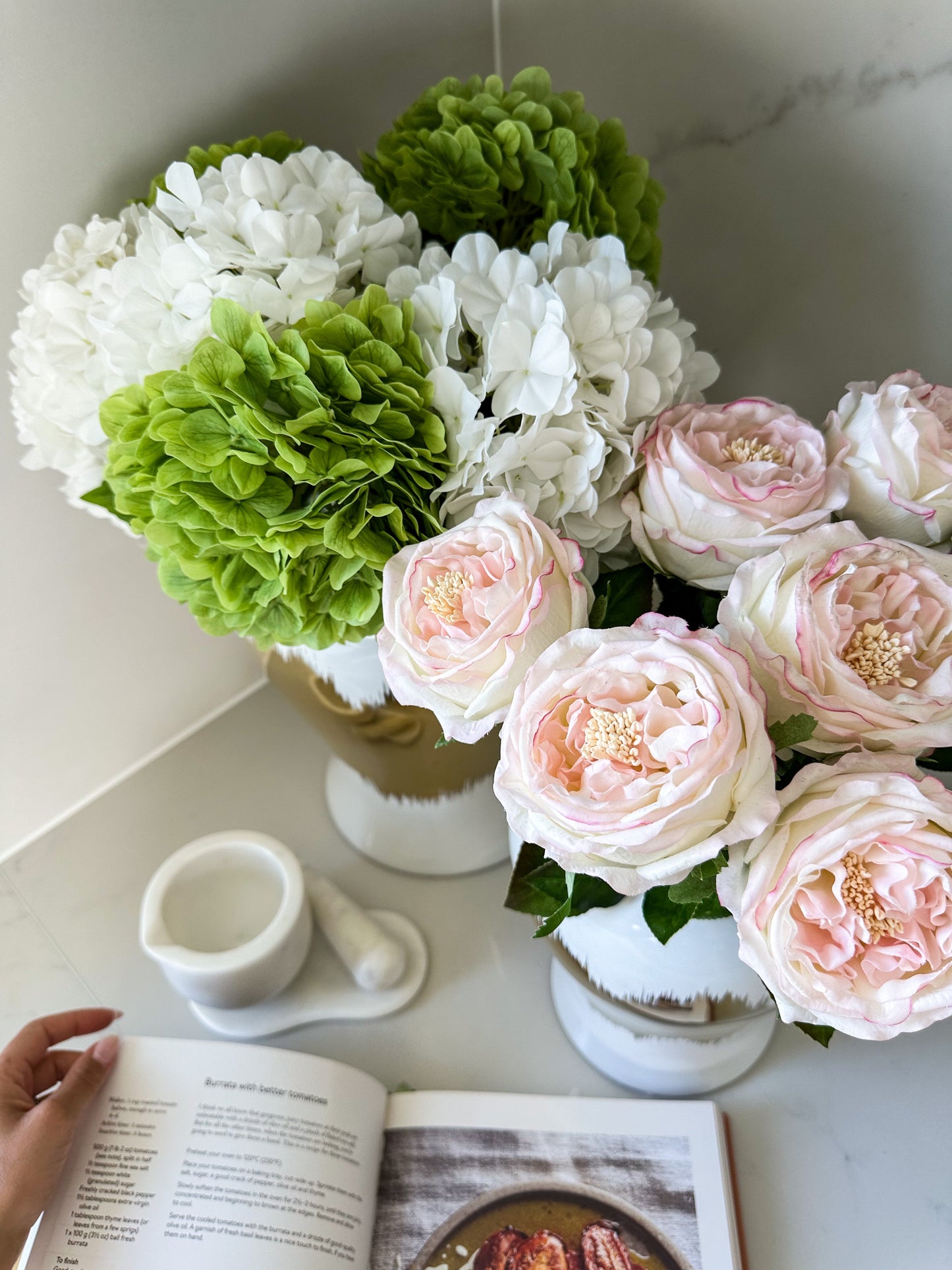 Top angled view of a collection of green and white real touch hydrangeas and light pink peonies on kitchen bench with recipe book