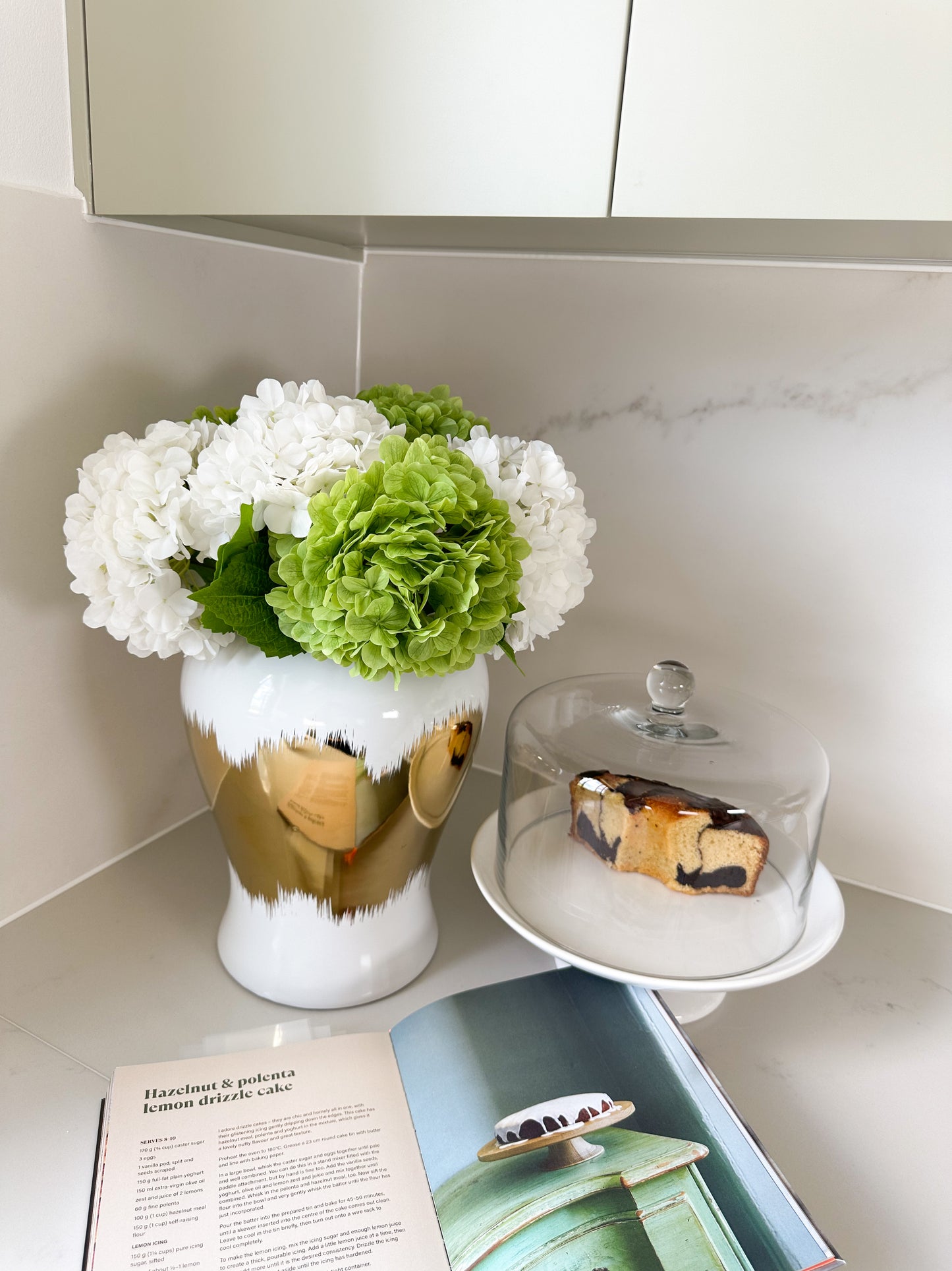 White and gold vase medium size with real touch hydrangeas in white and green on kitchen bench corner with recipe book in front of it 